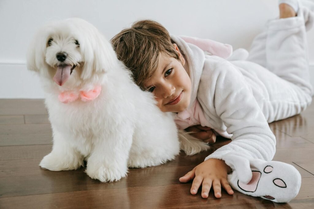 A child in cozy pajamas hugs a fluffy white Maltese dog, both lying on the wooden floor indoors.