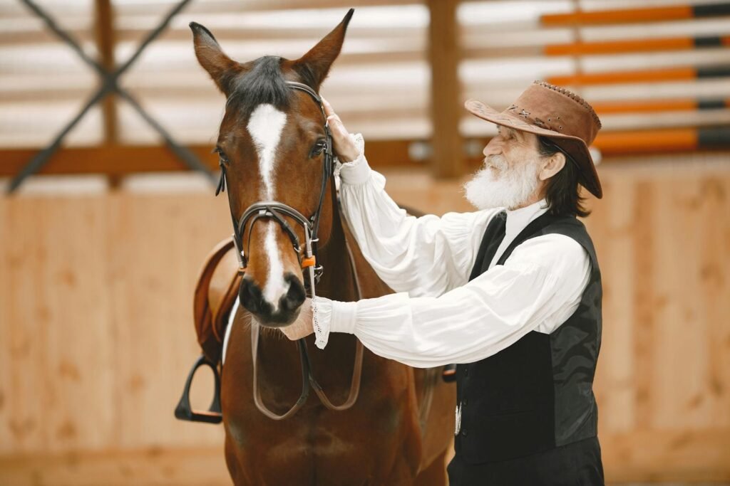 Elderly man with a beard and cowboy hat petting a horse in a stable.